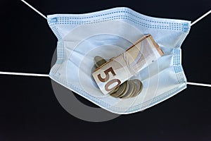 Medical face mask holding money inside of it, on a black background. World coronavirus epidemic and economic losses concept. Coins