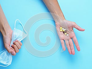 A medical mask is clutched in one hand and medications are lying in the other hand on a blue background.