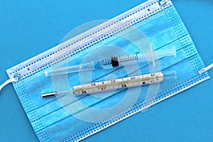 On a medical mask lie a thermometer and a syringe with medicine on a blue background.