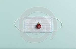 On the medical mask lies a New Year`s red ball imitating a sore red nose. Minimalistic flat lay on mint background