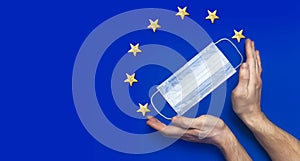 Medical mask in male hands on blue background flat lay top view. Flag of the European Union. Protection against virus, coronavirus