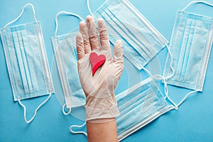 medical protective ear-loop masks and doctor`s hands in surgical gloves holding red wooden heart on blue background