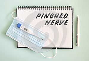 A medical protective mask and a thermometer are on the notebook. PINCHED NERVE text in a notebook