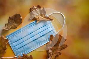 A medical protective mask and yellow dry oak leaves lie on a bright autumn background.