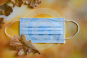 A medical protective mask and yellow dry oak leaves lie on a bright autumn background.