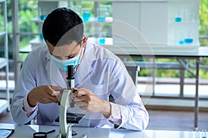 Medical scientist wearing protective surgical mask and lab gown using microscope in clinical laboratory