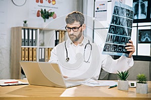 Medical specialist sitting at desk with x ray scan in hands and searching information in laptop