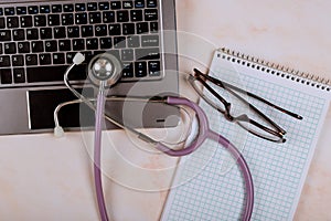 Medical stethoscope on laptop computer keyboard with blank notebook and glasses, working desk