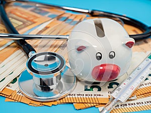 Medical stethoscope, piggy bank, thermometer and banknotes on a blue background.