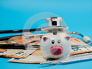 Medical stethoscope, piggy bank, thermometer and banknotes on a blue background.