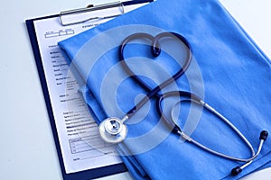 Medical stethoscope twisted in heart shape lying on patient medical history list and blue doctor uniform closeup