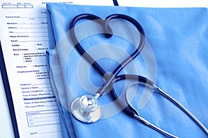 Medical stethoscope twisted in heart shape lying on patient medical history list and blue doctor uniform closeup