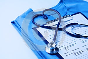 Medical stethoscope twisted in heart shape lying on patient medical history list and blue doctor uniform closeup