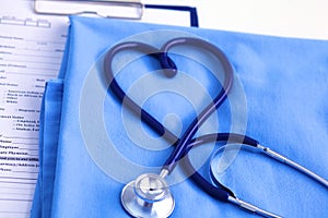 Medical stethoscope twisted in heart shape lying on patient medical history list and blue doctor uniform closeup