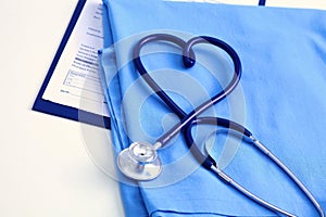 Medical stethoscope twisted in heart shape lying on patient medical history list and blue doctor uniform closeup