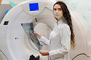 Medical student looks in camera with x-ray picture of patient brain in her hands. woman stays in front of kt komputer