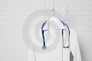 Medical uniform and stethoscope hanging on rack near white brick wall. Space for text