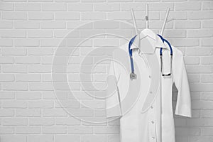 Medical uniform and stethoscope hanging on rack near white brick wall. Space for text