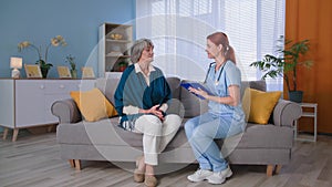 medical visit at home, young woman nurse in uniform with clipboard consulting senior female patient on sofa