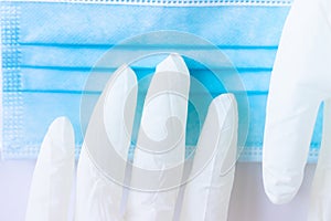 Medical white latex pair of gloves as a hand reaches for a blue mask on a white background.