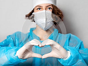 Medical worker in a disposable mask and a lab coat makes a heart sign. Gray homogeneous background. Conceptual photo: doctors on