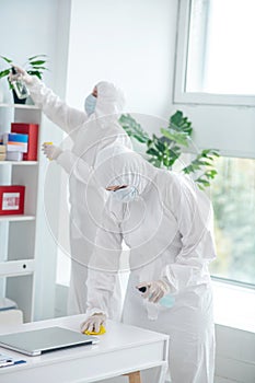 Medical worker in protective clothing and medical mask disinfecting table, another worker cleaning closet photo