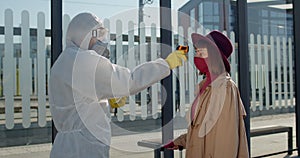 Medical worker in protective coverall suit and mask using tablet and screening female passenger while gesturing