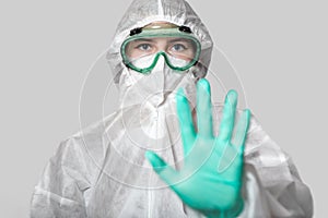 A medical worker in a protective suit, glasses, a respirator shows a STOP sign with his hand. Green latex gloves and face shield.