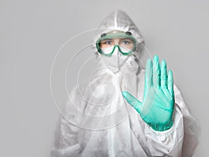 A medical worker in a protective suit, glasses, a respirator shows a STOP sign with his hand. Green latex gloves and face shield.