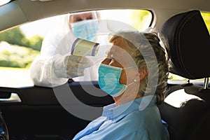 Medical worker wearing ppe suit taking temperature of senior caucasian woman sitting in car