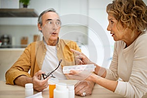 Medication supplements and treatment concept. Senior wife giving daily pills to her husband, sitting in kitchen interior