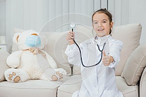 Little girl in medical coat holding a stethoscope, pointing her finger up. White teddy-bear in medical mask is sitting photo