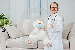 Pretty smiling little girl wearing doctor coat, stethoscope and glasses, standing, holding hand on the head of teddy photo