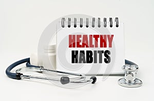 On a white table lies a stethoscope, pills and a notebook with the inscription - HEALTHY HABITS