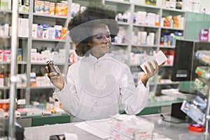 Medicine, pharmacy, healthcare concept. Happy female African apothecary worker posing at pharmacy holding two different
