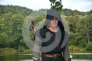 Medieval archer with black hood and arrows in the quiver stands before a lake and looks forwards