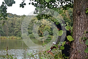 Medieval archer with black hood stands hidden behind tree in the lake with tense curve
