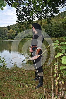 Medieval archer with black hood stands with tense curve and arrow on water