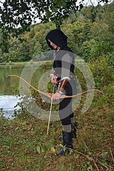 Medieval archer with black hood stands with tense curve and arrow on water