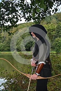 Medieval archer with black hood stands with tense curve and arrow on water