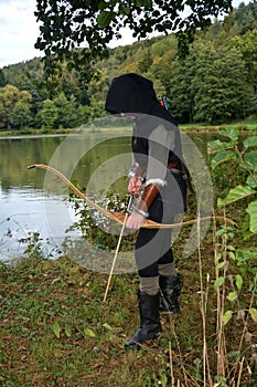 Medieval archer with black hood stands with tense curve and arrow on water