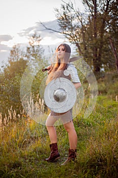 A medieval female warrior dressed in chain mail with a sword and shield in her hands poses against the background of a forest.