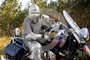 A medieval knight in chain mail and a helmet sits on a motorcycle.