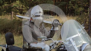 A medieval knight in full armor sits on a motorcycle against the backdrop of the forest