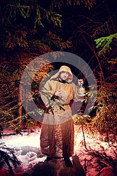 Medieval monk in canvas sackcloth robe with lattern in dark forest with snow and red light on winter night. Fantasy or fairy tale