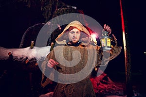 Medieval monk in canvas sackcloth robe with lattern in dark forest with snow and red light on winter night. Fantasy or fairy tale