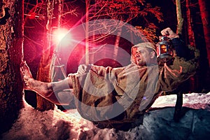 Medieval monk in canvas sackcloth robe with lattern resting on snow in dark forest and red light on winter night. Fantasy or fairy