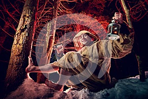 Medieval monk in canvas sackcloth robe with lattern resting on snow in dark forest and red light on winter night. Fantasy or fairy