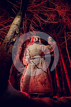 Medieval monk in canvas sackcloth robe with sword and lattern in dark forest and red light of moon on winter night. Fantasy or