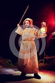 Medieval monk in canvas sackcloth robe with sword and lattern in dark forest and red light of moon on winter night. Fantasy or